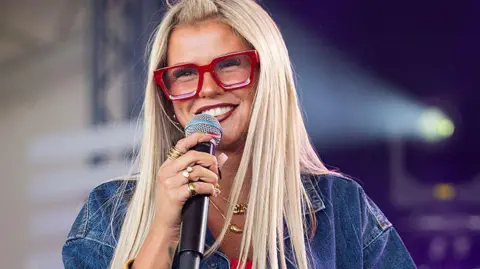 Getty Images Skye is performing in a dark blue denim jacket and wearing wide-rimmed red glasses. She has long blonde hair and has lots of gold rings on her fingers and two necklaces. She is smiling and wearing dark red lipstick.