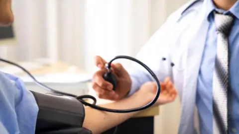 Getty Images Doctor in white coat, with blue shirt and grey tie takes blood pressure from a patient wearing a blue top.