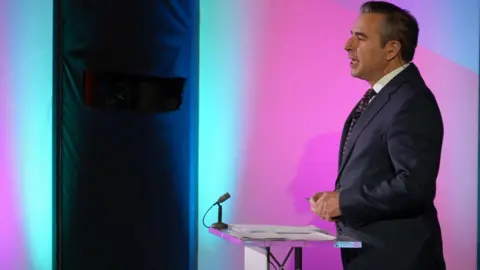 Nick Servini standing at a lectern and speaking, wearing a dark suit and tie. Behind him is a purple and blue set. The lectern has two small piles of notes on it and a small microphone on one of two corners of it furthest away from Nick.