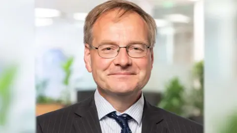 Head and shoulders of Malcolm Johnson in a grey suit, blue tie and is wearing spectacles