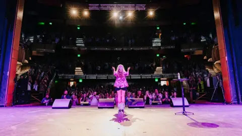 Serena Terry images A picture from behind Ms Terry on stage. The audience are facing the camera but are in slight darkness. Ms Terry is facing them, wearing a pink jacket and she is standing in the spotlight. 