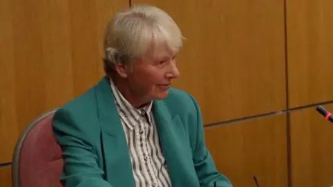 Qays Najm/BBC Rosemary Thew sits at a desk inside the council chamber. She is wearing a green jacket, striped black and white shirt and has white hair.