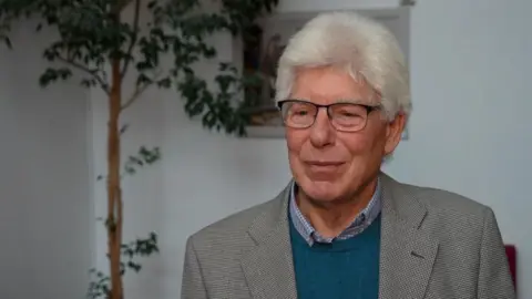 Qays Najm/BBC Councillor Paul Heinrich has white hair, wears glasses and is wearing a blue and white shirt, turquoise jersey and checked jacket. He is in a room painted white, with a potted tree in the background.