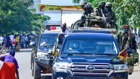 State House Uganda/X Soldiers with guns at the back of a black truck and others leaning out of the doors as a convoy goes down a street in Uganda.