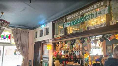 Hayley Coyle/BBC Interior of a cozy pub or bar with a decorated counter and hanging scarves. Visible scarves include ones labeled 'ARCTIC MONKEYS' and 'IRELAND.' Signs read 'TUMBLING PADDIES-25' and 'CORK.' The bar is adorned with bottles, glasses, flowers, and various decorations, with stained glass windows and curtains in the background.