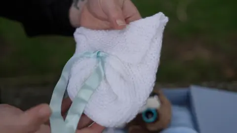 BBC/Joe Weir  A small white knitted garment with a green ribbon is held by female hands.