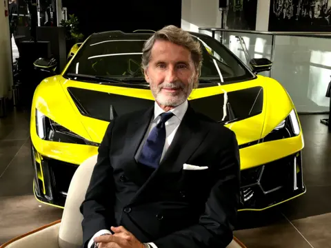Lamborghini chief executive Stephan Winkelmann in a full suit, sat in front of a bright yellow Lamborghini Fenomeno