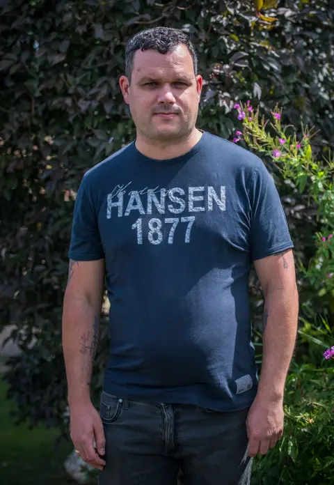 Ben, the father of Alice's eldest daughter Delsie.  He is wearing a blue jeans and a t-shirt with Hansen 1877 logo on it.  He as short dark brown hair. 