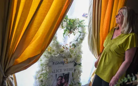 Alice Webb's mother Rachael stands next to a window and a funeral floral arrangement with photos of Alice. 