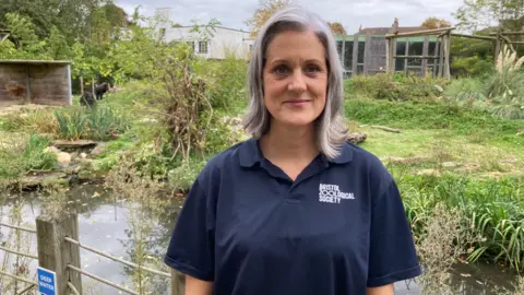 Hannah Windross. She has grey hair and is wearing a blue t-shirt with the word "bristol zoological society" on it. She is standing in front of the gorilla enclosure