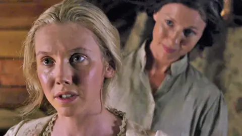 Starz TV A still from Outlander shows a young blonde woman in period costume - wearing a light-coloured dress with lace trim - she is closer to the camera and more prominently featured. In the Background is an older woman dressed in a darker, plain dress. They are in a wooden panelled room.