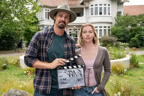 BBC/The Ridge Production Ltd A behind-the-scenes moment from The Ridge shows Lauren Lyle and Jay Ryan   standing in front of a large white house with a red-tiled roof and multiple windows. Ryan wears  a hat, a checked shirt over a green t-shirt, and holding a clapperboard used in film production. Lauren Lyle is dressed in a light purple top, an open cardigan, and jeans. 