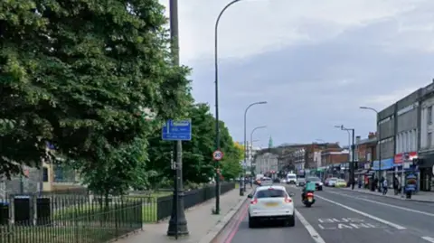 Google Google Maps shot of a Catford street called Rushey Green showing cars and a motorbike in a bus lane. There pavement to the left is lined with trees and contains two patches of grass surrounded by iron fencing.