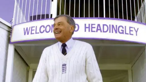Reuters Veteran cricket umpire Harold "Dickie" Bird walks onto the pitch at Headingley in Leeds before umpiring his last ever cricket match between Yorkshire and Warwickshire September 13. He is wearing a white sweater with an emblem, a collared shirt, and dark pants stands in front of a building entrance with a sign that reads 'WELCOME TO HEADINGLEY.' 