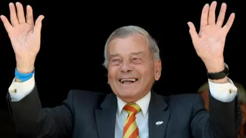 Anthony Devlin/PA Wire Dickie Bird, an elderly man with short grey hair, dressed in a dark suit, white shirt, and a striped yellow and red tie. He is smiling broadly and raising both hands in the air. The background is dark, with other people partially visible behind.
