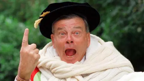 Paul Barker/PA Wire Dickie Bird: a man wearing a black hat with a yellow tassel and a large white scarf around their neck has their mouth open and eyes wide, with one hand raised and pointing upward. The background features blurred greenery.