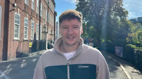 Brett wearing a cream jumper smiling on campus