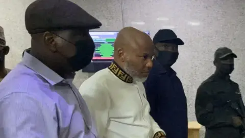 Getty Images Nigerian separatist leader Nnamdi Kanu dressed in white with three nose-masked Nigerian security officers by his side.