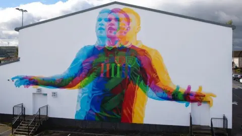 Gasyard Féile A mural on a white wall of footballer James McClean - he is wearing a Republic of Ireland kit and his his arms outstretched