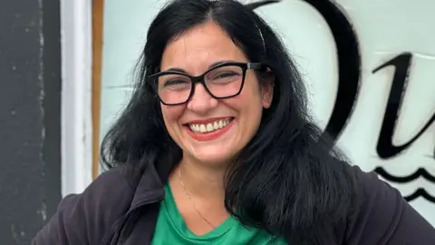 Matilde Liso, who has long dark hair and is wearing black rimmed glasses and a black top over a green T-shirt. She is smiling.