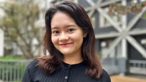 Audrey Hill-Uekawa, 20,  is wearing a black cardigan and has shoulder lenght black hair. She is smiling for the camera standing outside in front of a tree and a building