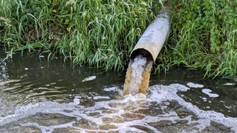 Getty Images An overflow pipe discharges a brown liquid into a stream or river. 