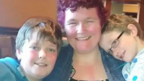 Supplied Claire Throssell smiles as she hugs her children Jack and Paul. She has short hair, dyed purple, and wears a blue denim shirt. Jack has brown hair, wears a blue top and is smiling into the camera. Paul has light-brown hair and wears glasses and a white and blue top.