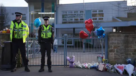 PA Media Police stand at the gate of the school. Next to them are flowers, notes and balloons.
