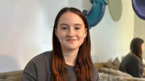 Grace sits on a couch and smiles at the camera. She has long brown hair.