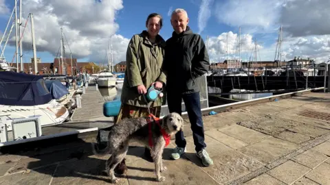 Kevin Shoesmith/BBC A middle-aged couple are pictured with their grey lurcher dog, with boats moored on a marina behind them. The woman is wearing a long, olive green jacket. Her husband is wearing a black jacket and navy jeans.  