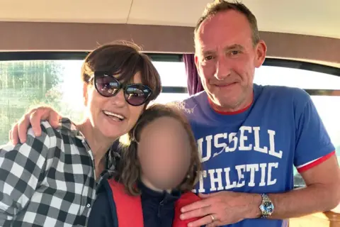 Marie Fuller A woman with short dark brown hair, wearing black sunglasses and a black and white checked shirt is hugging a young girl, also with shoulder-length brown hair. The girl, whose face has been blurred, is wearing a dark top and and a red life vest. On the right hand side of her is a man with receding hair and a blue t-shirt which says Russell Athletic on it.