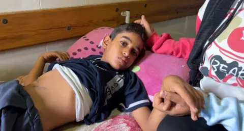 A Palestinian boy, Ahmed al-Jadd, lies on a bed, looking poorly and holding a woman's hand
