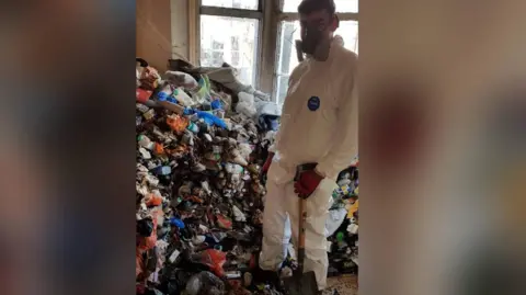 Ben Giles Photograph shows a picture of a pile of rubbish mounted up in the bay of a window. A boy stands in front of the pile of rubbish. He wears full PPE, a face mask and holds a shovel. He wears black and red gloves.