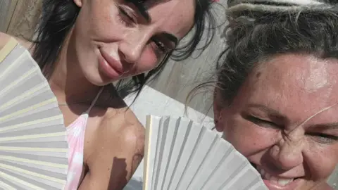 Handout Beth Ashton, who has long dark hair, poses for a picture with her mother Natalie Tolan, who has blonde hair. They are both holding paper fans. 