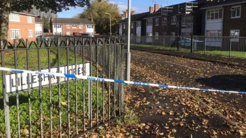 A road with two-storey houses on either side behind a black fence. A white signs with black text that reads "Lilleshall Crescent". There is blue and white police tape across the road and lots of orange leaves on the ground.