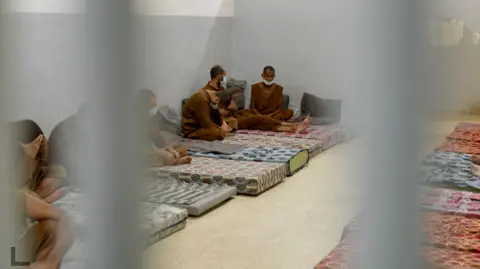 Goktay Koraltan/BBC Several detainees in brown uniforms with shaven heads sitting on mattresses inside  al-Sina prison. 