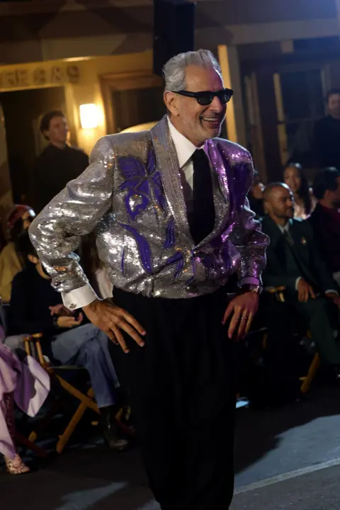 Getty Images Jess Goldblum wearing a silver jacket and sunglasses stands with hands on hips, smiling. 