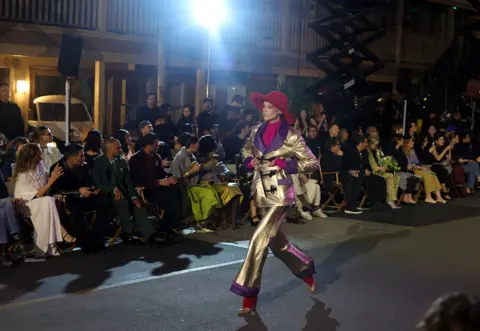 Getty Images A model strides along the runway during Vogue World: Hollywood 2025 at Paramount Studios in a gold suit with flaired trousers, purple trim and fushia pink blouse and socks. The audience claps in the background