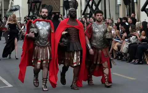 Getty Images Three male performers in  red and brown gladiator costumes walk next to each other along the runway during Vogue World: Hollywood 
