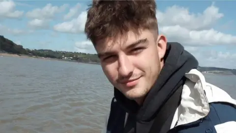 Family photo A young man with short dark hair, with a black scarf around his neck and a white jacket. He is standing in front of a body of water.