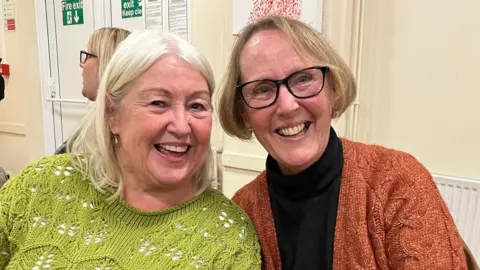 Two middle-aged women in jumpers, leaning their heads towards each other for the photo, both smiling broadly.