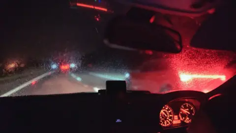 Getty Images/Stephen Robinson Pictures The driver's view out of the front of a car on a dark, rainy night. Inside the car dials on the dashboard are lit up and the rear-view mirror is visible. Through the windscreen you can see blurred oncoming headlights in the distance.