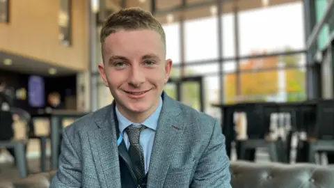 Fraser wears a grey blazer with a blue shirt and grey tie. He is sitting in the school theatre bar with floor to ceiling glass windows, you can see tables and chairs behind him. He is smiling at camera.
