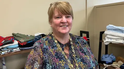 Deputy Mayor Niree McMorris is wearing her gold chain of office. She's standing in a room with baby and children's clothes folded on shelves. 