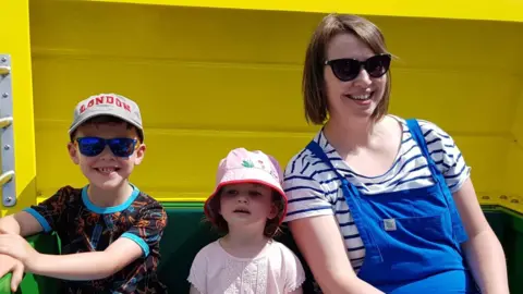 Darren Wheatley Nicola with Oscar and Ffion. The children are wearing sunhats and t-shirts and Nicola is wearing blue dungarees over a blue and white striped t-shirt. All are smiling. 