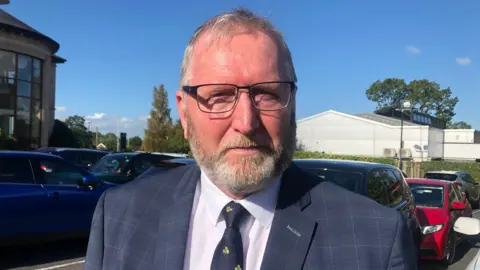 Doug Beattie is wearing a navy blazer and white shirt with tie. He has glasses on. 