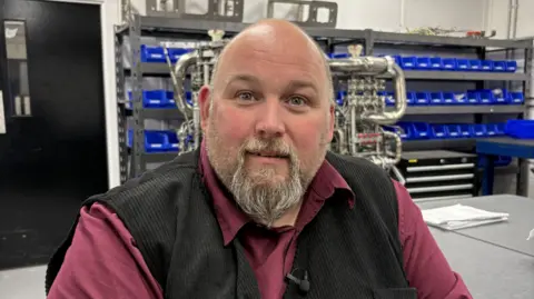Derek Harris  fom Skyrora, a bald man with a light coloured bears, wearing a black waistcoat and a burgyndy shirt. He is smiling. Behind him are shelves with rows of blue containers and a work table with a metal structure on it.