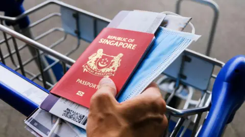 Getty Images In airport: hand on luggage trolley, holding Singapore passport, boarding pass, face mask, and map. 