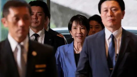 Reuters Japan's new Prime Minister Sanae Takaichi arrives at the prime minister's official residence in Tokyo, Japan October 21, 2025. She is wearing a blue suit, with a pearl necklace and earings. Her hair is short. She is surrounded on all sides by men in suits
