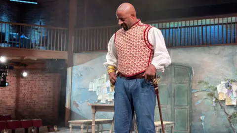 BBC A man standing on a stage during a performance of a play. He has an enhanced, disfigured nose, a red and white waistcoat over a white shirt, a sheathed sword, jeans and is looking down with his hands at his pockets. Behind him are tables and chairs, as well as a balcony.